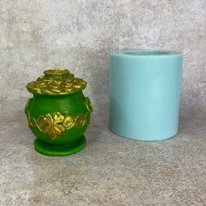 May include: A green pot of gold candle mould with a gold coin design and a shamrock. The mould is sitting on a light blue surface next to a cylindrical silicone mould.