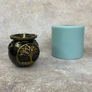 May include: A gold candle with a tree of life design in a black pot. The candle is next to a light blue silicone mould.