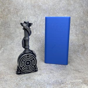 Puede incluir: Un portavelas gris oscuro detallado con una serpiente enrollada alrededor del tallo y un diseño laberíntico en la base. Un objeto rectangular azul está a la derecha del portavelas. El portavelas está sobre una superficie gris.