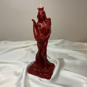 May include: A red wax candle sculpted in the shape of a woman wearing a crown and holding a cornucopia. The candle is standing on a square base.