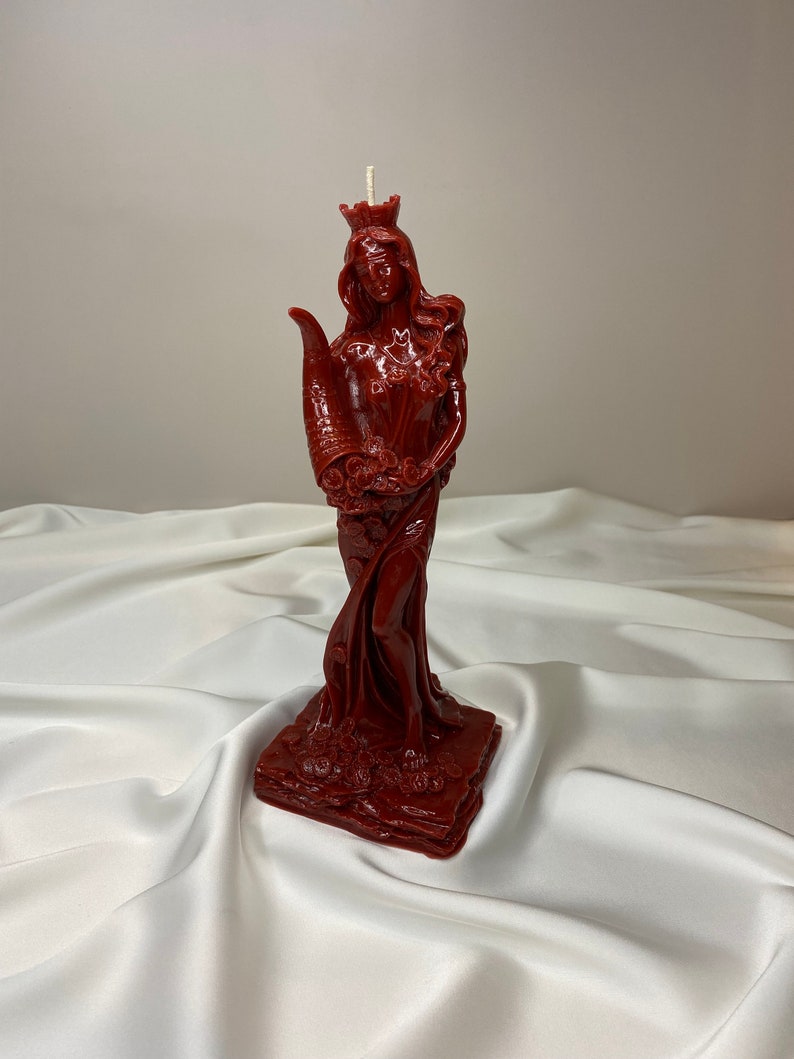 May include: A red wax candle sculpted in the shape of a woman wearing a crown and holding a cornucopia. The candle is standing on a square base.