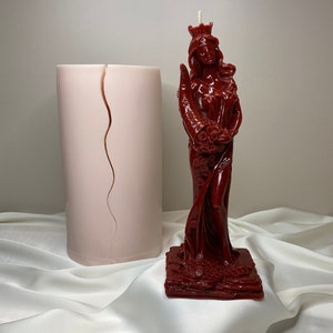 May include: A red candle in the shape of a woman wearing a crown and holding a bouquet of flowers. The candle is on a square base and is surrounded by a white fabric.