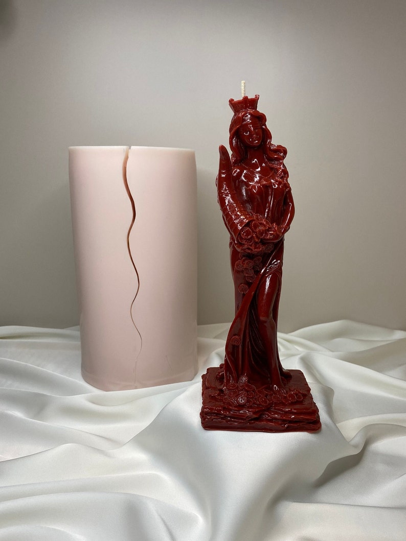 May include: A red candle in the shape of a woman wearing a crown and holding a bouquet of flowers. The candle is on a square base and is surrounded by a white fabric.