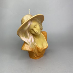 Può includere: Una candela dorata e beige a forma di busto di donna con un cappello a tesa larga. La candela ha un viso e capelli dettagliati.