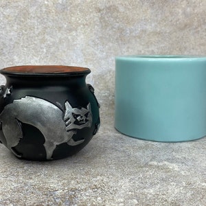 May include: A black cauldron-shaped ceramic pot with a silver cat design and a light blue cylindrical container. The cauldron has a brown interior and is decorated with stars. The cat has an angry expression.