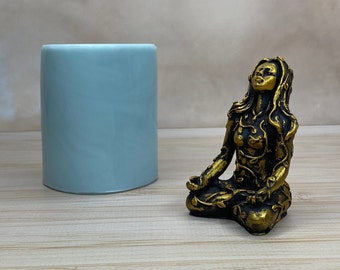 May include: A gold-colored figurine of a woman sitting in a meditative pose. She is covered in leaves and vines, and her eyes are closed. The figurine is sitting on a light wood surface.