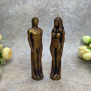 Puede incluir: Dos velas de color dorado con forma de figuras humanas. Las velas están de pie sobre una superficie texturizada.
