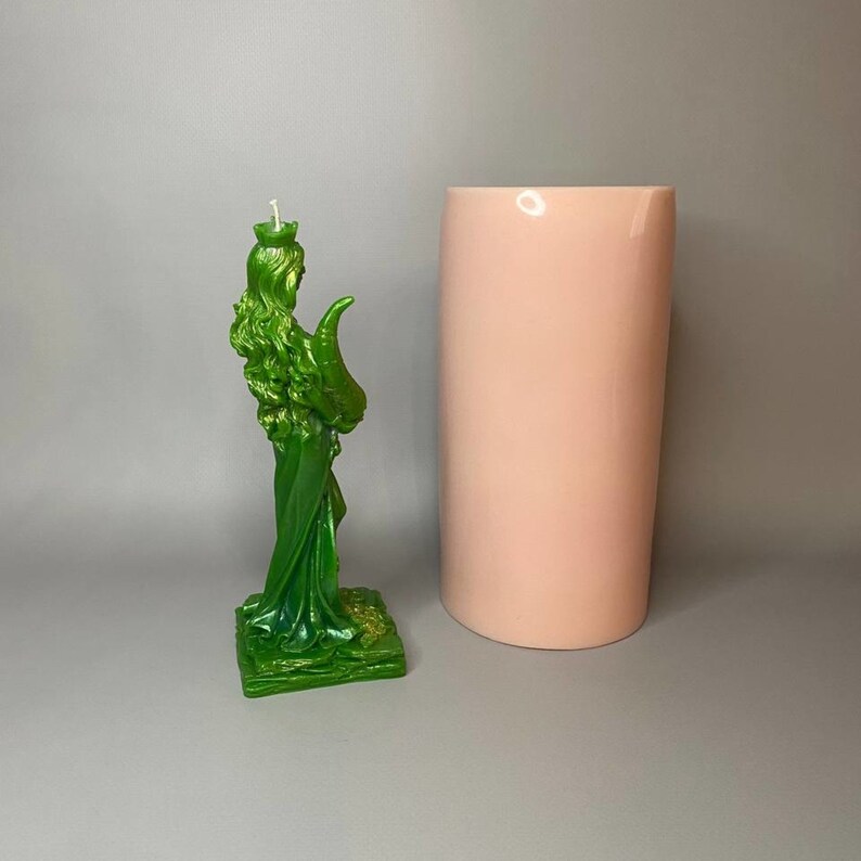 May include: A green candle shaped like a woman holding a horn. The candle is on a green base and has a white wick. The candle is next to a pink cylindrical candle.