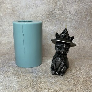 May include: A black silicone mold for making a candle in the shape of a black cat wearing a witch's hat with a pentagram and skull accents. The mold is next to a finished candle.