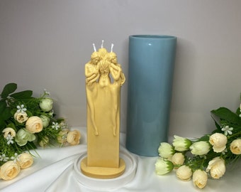 May include: A yellow candle shaped like three women standing together. The candle is on a white base and has three wicks.