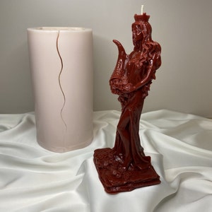 May include: A red wax candle sculpted in the shape of a woman with a crown and flowers. The candle is standing on a red base with floral details. A white cylindrical candle mold is in the background.