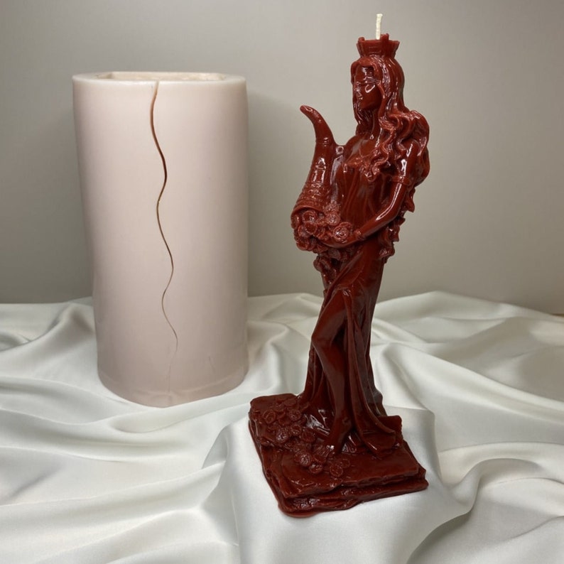 May include: A red wax candle sculpted in the shape of a woman with a crown and flowers. The candle is standing on a red base with floral details. A white cylindrical candle mold is in the background.