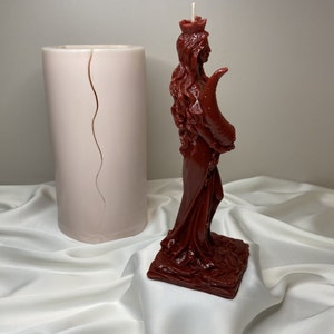 May include: A red, detailed candle in the shape of a woman wearing a crown and holding a crescent moon. The candle is on a square base and is sitting on a white fabric surface.