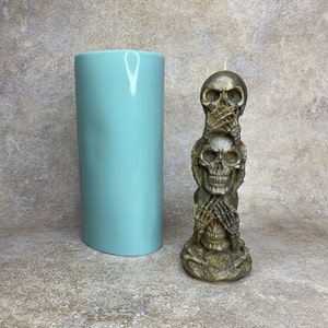 May include: A three-skull candle with a brown finish. The top skull has its hands over its mouth, the middle skull has its hands over its eyes, and the bottom skull has its hands over its ears. The candle is sitting on a light brown surface.