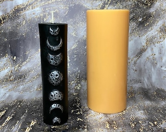 Puede incluir: Dos velas, una negra con un diseño de fases lunares plateadas y otra naranja lisa. La vela negra es más corta que la vela naranja.