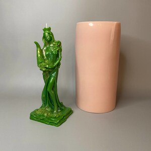 May include: A green candle in the shape of a woman holding a cornucopia. The candle is on a green base and is next to a pink cylinder.