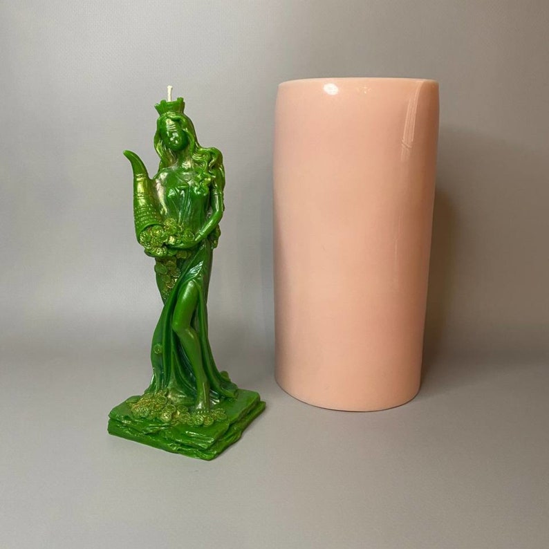 May include: A green candle in the shape of a woman holding a cornucopia. The candle is on a green base and is next to a pink cylinder.
