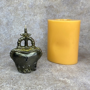 May include: A decorative candle in the shape of a heart with a crown design, in antique gold and dark gray. Next to it is a cylindrical, unscented candle in a light yellow color. The candle has a wick.