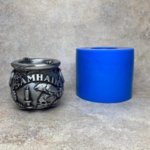 May include: A silver cauldron with the word "SAMHAIN" embossed on the front, featuring a candle, raven, and skull design. A bright blue cylindrical mould is next to the cauldron. The cauldron is likely used for crafting or decoration.