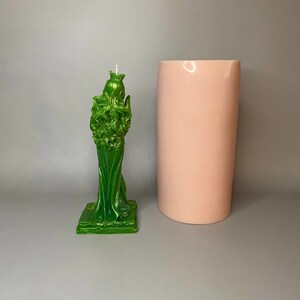 May include: Green candle in the shape of a woman wearing a crown and a flowing robe. The candle is on a square base. A pink cylindrical mold is next to the candle.