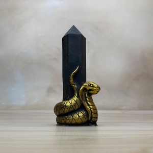 May include: A black obsidian pyramid with a gold snake wrapped around it. The snake is coiled around the base of the pyramid and its head is facing forward.