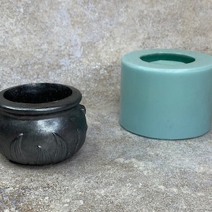 May include: A black cauldron-shaped silicone mould for making candles or soap. The mould is sitting on a light grey surface next to a finished black cauldron.