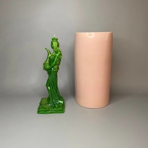May include: A green candle in the shape of a woman holding a horn. The candle is on a green base and is standing next to a pink cylinder.