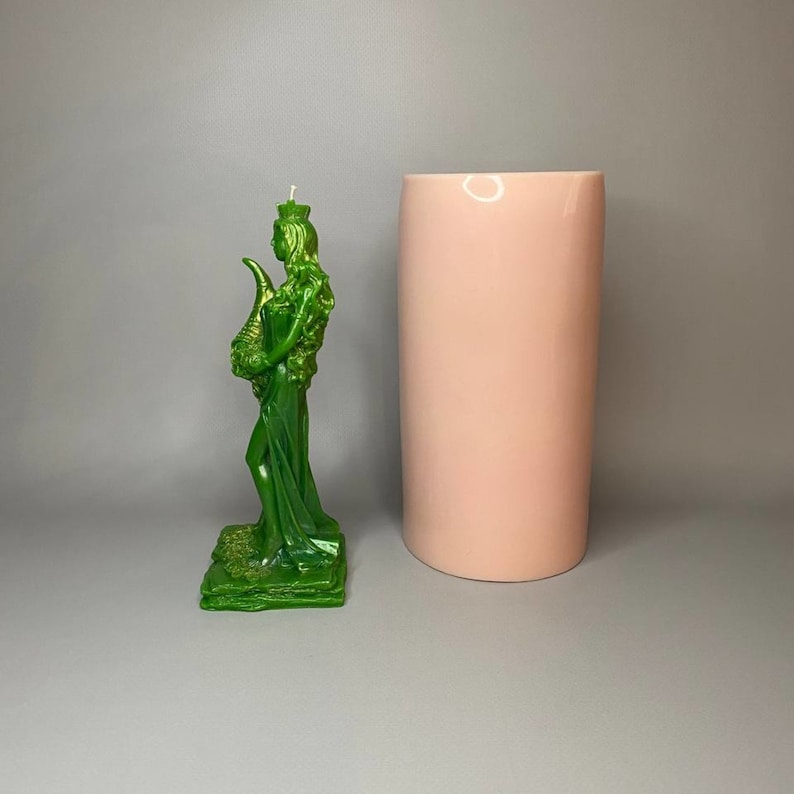 May include: A green candle in the shape of a woman holding a horn. The candle is on a green base and is standing next to a pink cylinder.