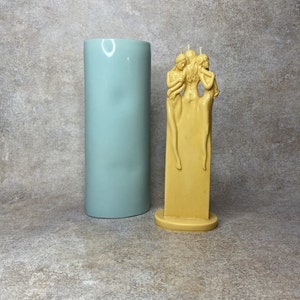 Pu&ograve; includere: Una scultura di candela a tre figure di donne in piedi vicine. La candela &egrave; di colore giallo pallido e si trova su una piccola base rotonda. La candela &egrave; accanto a uno stampo cilindrico blu chiaro.