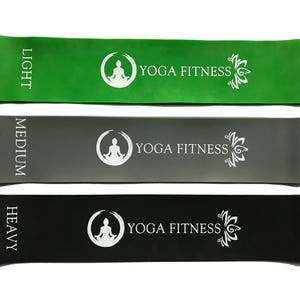 Puede incluir: Tres bandas de resistencia en diferentes colores y niveles de resistencia: verde claro, gris medio y negro intenso. Cada banda tiene el texto "YOGA FITNESS" y una figura de yoga estilizada impresa en ella.