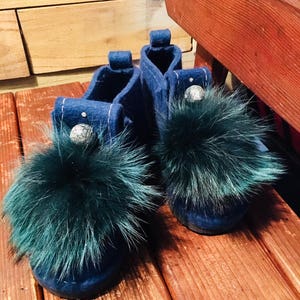 May include: A pair of blue felt slippers with green fur pom-poms. The slippers have a decorative silver bead on each.