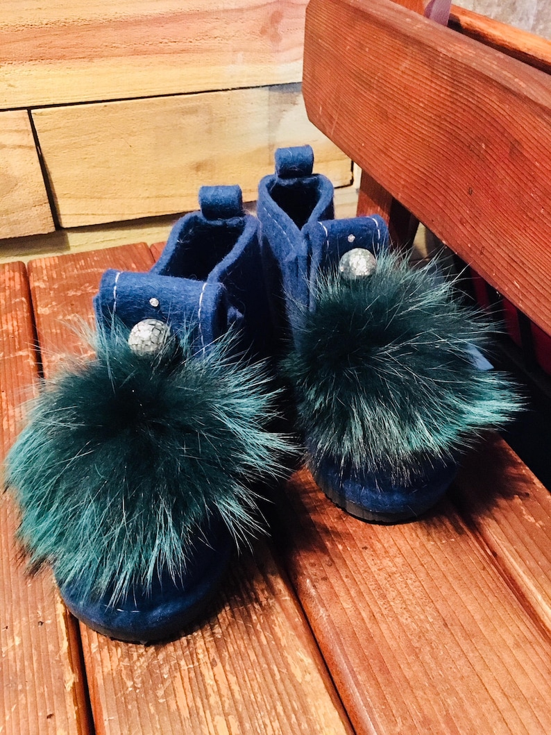 May include: A pair of blue felt slippers with green fur pom-poms. The slippers have a decorative silver bead on each.