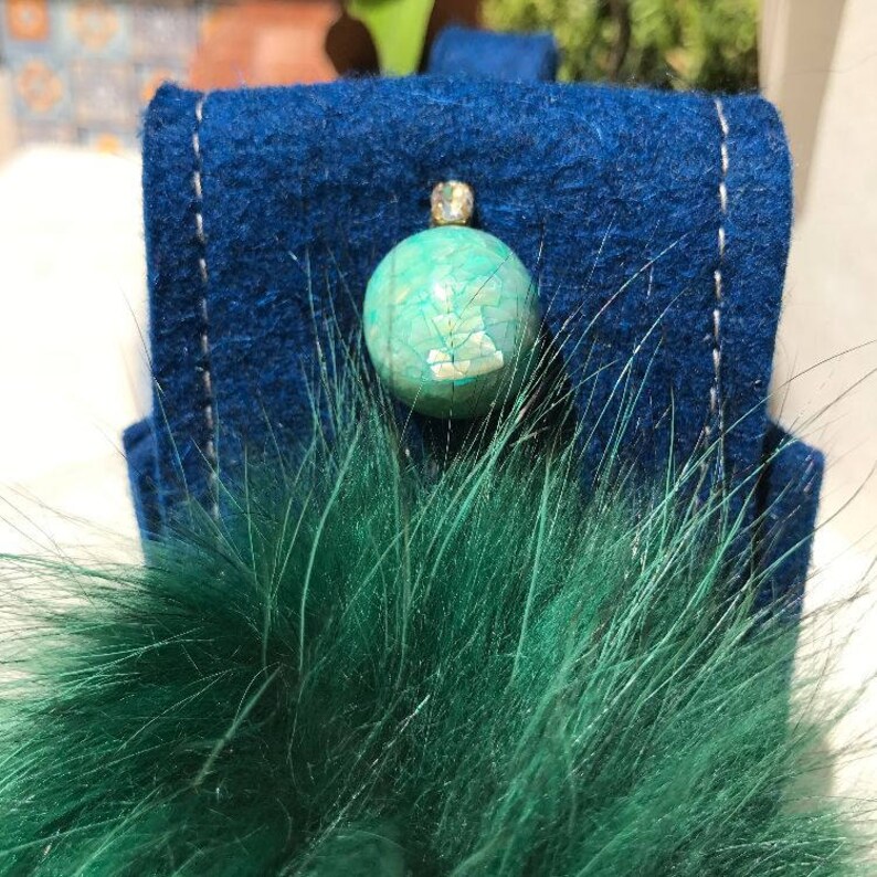 May include: A blue felt bag with a green fur pom-pom and a turquoise gemstone embellishment.