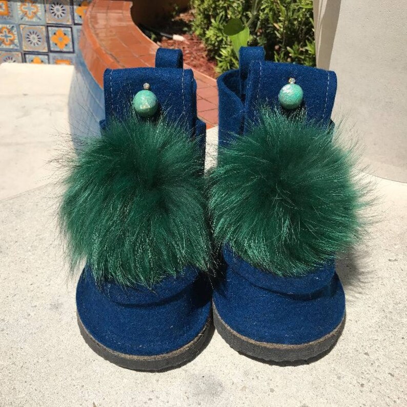 May include: A pair of dark blue felt boots with green fur pom-poms and turquoise accents. The boots have a rounded toe and a flat sole.