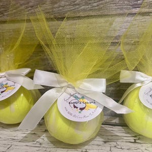 Puede incluir: Varias bombas de baño redondas y amarillas están envueltas en tul amarillo y atadas con cintas de satén blancas. Cada bomba tiene una pequeña etiqueta redonda con el texto "Southern Banana Cabana".
