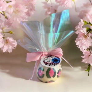 May include: A round, cream-colored product with a pink and green leopard print design, wrapped in iridescent cellophane and tied with a pink ribbon. A small tag with text and a blue flower is attached. Pink flowers are in the background.