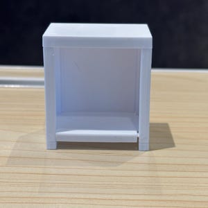 May include: A small, white, rectangular shelf unit with an open front and a shelf. The unit is made of a smooth, solid material and sits on a wooden surface. The design is simple and minimalist.