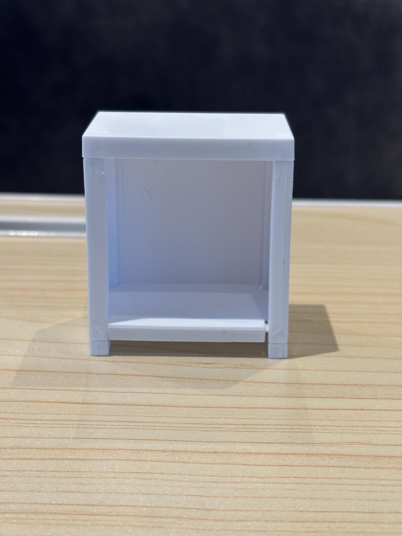 May include: A small, white, rectangular shelf unit with an open front and a shelf. The unit is made of a smooth, solid material and sits on a wooden surface. The design is simple and minimalist.