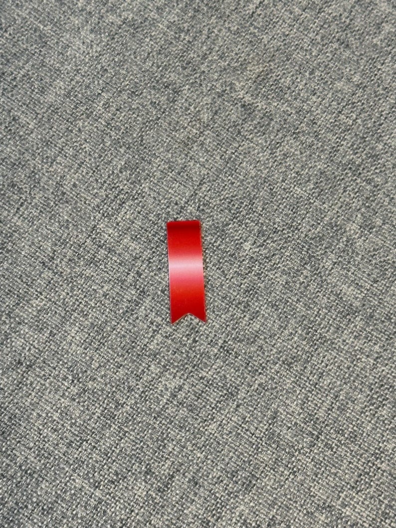 May include: A small, bright red ribbon with a pointed bottom edge. The ribbon is positioned vertically against a textured gray background, creating a simple yet striking contrast. The ribbon's color is vibrant and eye-catching.