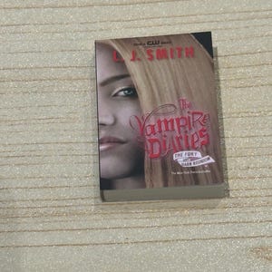 May include: A paperback book titled "The Vampire Diaries" by L.J. Smith. The cover features a woman's face with blonde hair and the title in red lettering. The book is a New York Times bestseller.