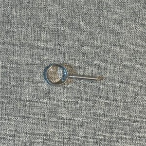 May include: A small, silver-toned magnifying glass with a clear, circular lens and a short handle. The magnifying glass is resting on a textured, gray surface.