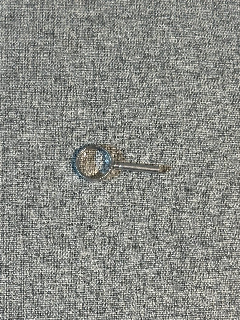 May include: A small, silver-toned magnifying glass with a clear, circular lens and a short handle. The magnifying glass is resting on a textured, gray surface.