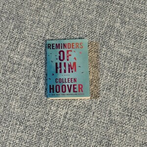 May include: A paperback book titled "Reminders of Him" by Colleen Hoover. The cover is a teal color with red lettering. The title is in large, bold letters, and the author's name is below. Small bird illustrations are scattered across the top.