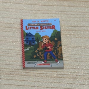 May include: A children's book titled "Baby-Sitters Little Sister" by Ann M. Martin. The cover features an illustration of a girl in a red shirt and blue pants, standing in front of a house. The book is published by Scholastic.