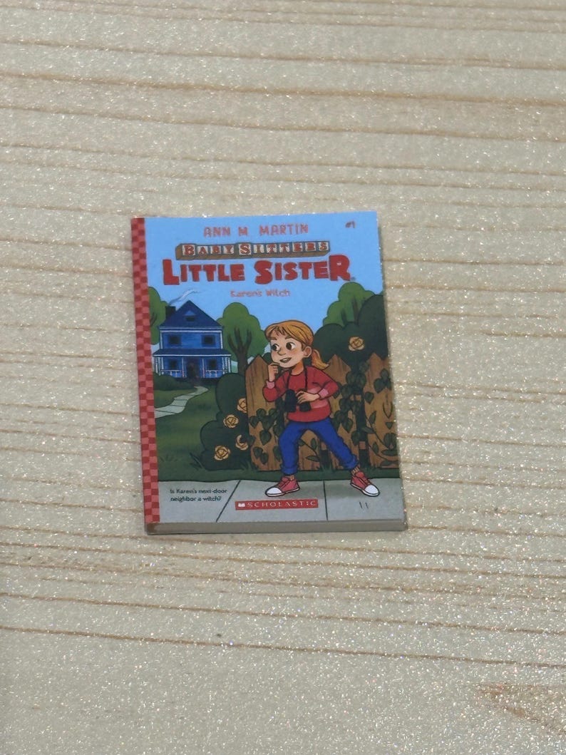 May include: A children's book titled "Baby-Sitters Little Sister" by Ann M. Martin. The cover features an illustration of a girl in a red shirt and blue pants, standing in front of a house. The book is published by Scholastic.