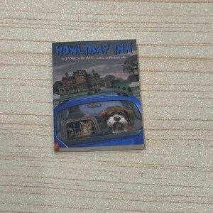 May include: A book titled "Howliday Inn" by James Howe. The cover features a blue car with a dog and a cat looking out the back window. A large house is in the background.