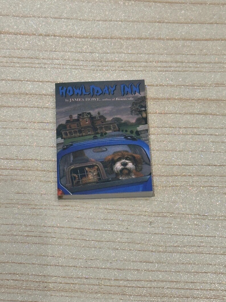May include: A book titled "Howliday Inn" by James Howe. The cover features a blue car with a dog and a cat looking out the back window. A large house is in the background.