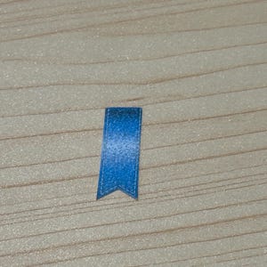 May include: A small, blue bookmark with a textured appearance. The bookmark has a pointed bottom edge and a stitched border. The background is a light-colored wooden surface.