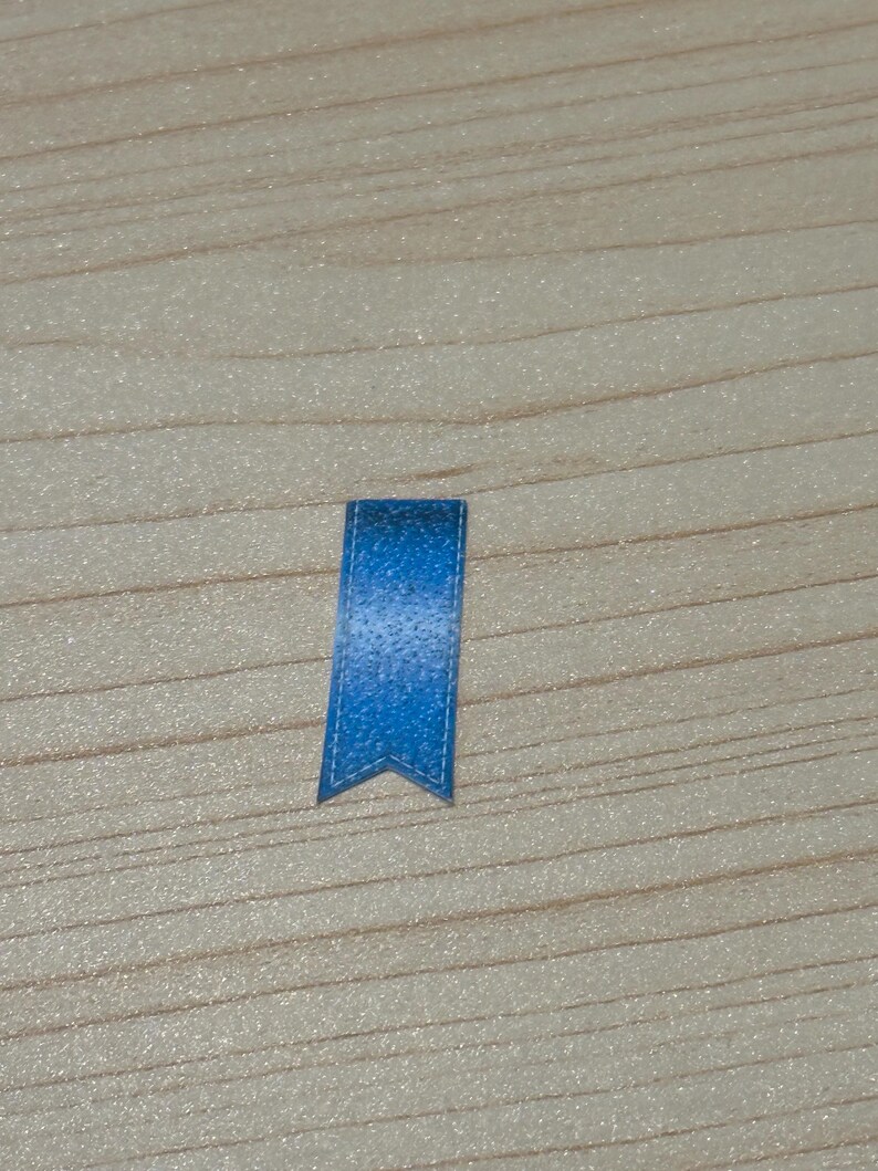 May include: A small, blue bookmark with a textured appearance. The bookmark has a pointed bottom edge and a stitched border. The background is a light-colored wooden surface.