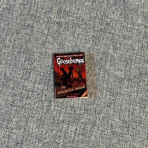 May include: A paperback book titled "Goosebumps: The Haunted Mask" by R.L. Stine. The cover features a dark red background with silhouetted figures and the title in a spooky font. The book is set against a gray textured surface.